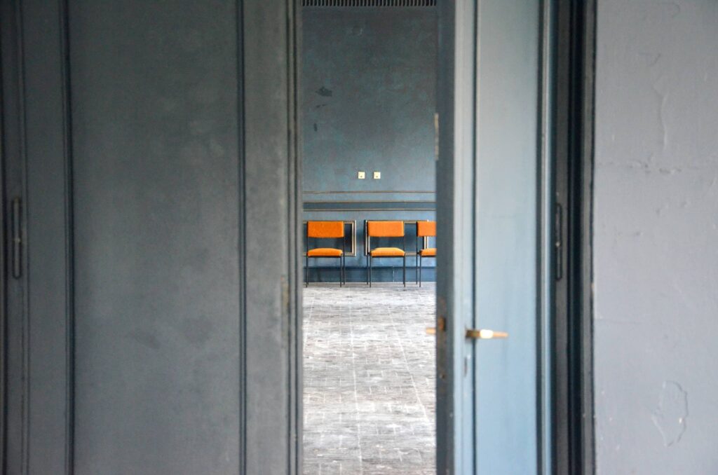 Aktuelle Innenaufnahme des Kulturhauses: Durch eine halb geöffnete Tür fällt der Blick in einen leeren Raum mit drei orangefarbenen Stühlen vor einer blaugrauen Wand. Die Szenerie vermittelt eine typische Lost-Place-Atmosphäre. 2025.